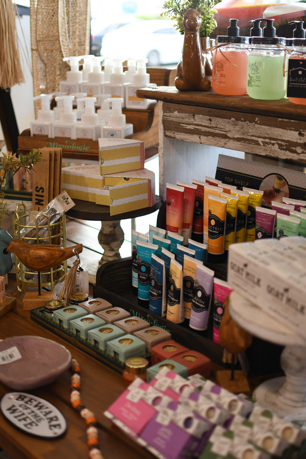 A retail display table filled with various handmade soap bars, tubes of body wash, and boxed products from brands like 'Sift & Infuse' and 'Bourgeon', set in a rustic shop environment.