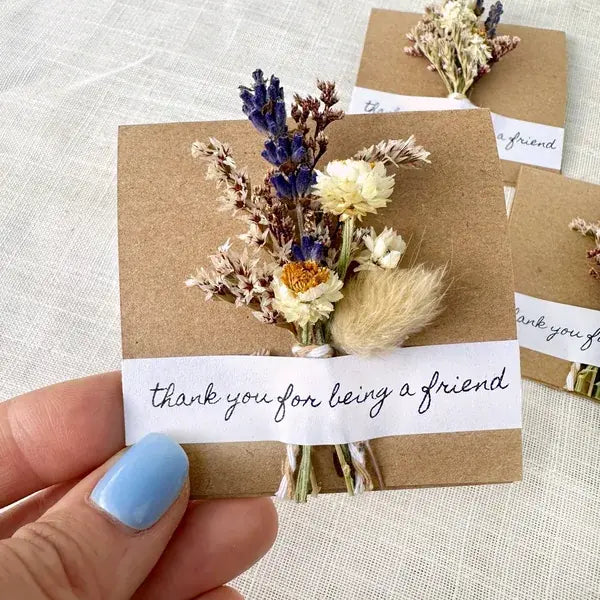 A hand with light blue nail polish holds a small dried flower card that says 'thank you for being a friend' on a white strip, with kraft paper backing and a bouquet of beige, cream, and purple dried flowers. Other similar cards are visible in the background on a textured off-white fabric surface.