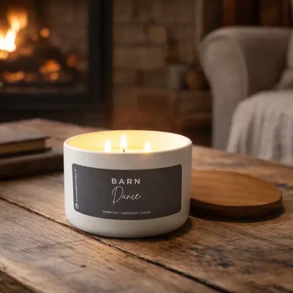 Candle with a label reading 'Barn Dance' on a wooden surface in front of a fireplace.