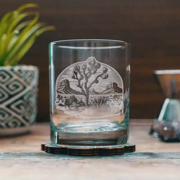 A clear glass rocks tumbler with an etched desert landscape design featuring a Joshua tree and rock formations, resting on a dark wooden coaster on a rustic wooden table.