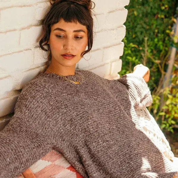 A woman with dark hair in a bun leans against a white brick wall, wearing a chunky knit crewneck sweater in a heathered taupe-brown color. A cream knit blanket is draped over her lap, and green foliage is visible in the background.