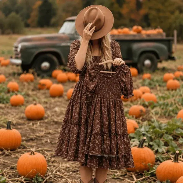 A woman stands in a pumpkin field wearing a brown floral tiered dress, a wide-brimmed brown hat, and holding a small wooden basket. A vintage green pickup truck loaded with pumpkins is in the background.