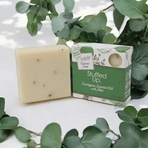 A bar of natural soap labeled 'Stuffed Up.' sits beside its matching green and white box, styled with eucalyptus leaves on a white surface.