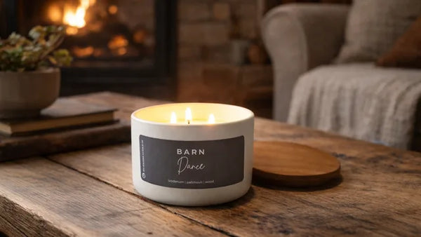 A lit white candle jar labeled 'BARN Dance' sits on a rustic wooden table in a cozy living room setting with a fireplace and sofa in the background.