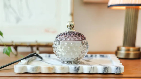 A textured glass jar with a matching glass lid and a small gold finial, displayed on a white marble tray on a wooden table in a softly lit interior setting.