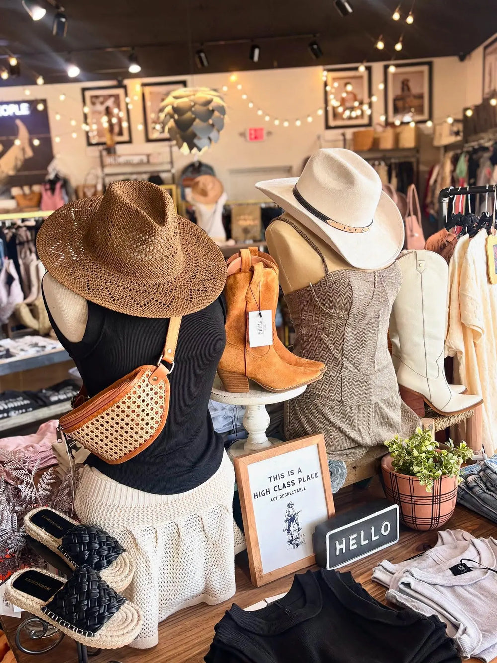 A boutique display featuring mannequins dressed in summer fashion, including a straw hat, black top, white skirt, tan cowboy boots, and a woven crossbody bag. The store interior is visible in the background with racks of clothing and string lights.