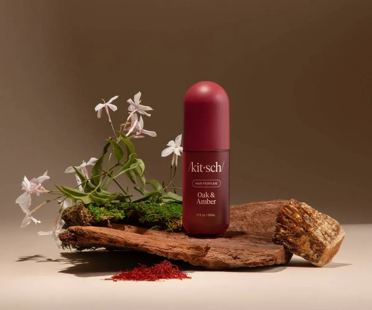A Kit-Sch Oak & Amber deodorant bottle with a dark red cap and label stands on a rustic wooden slab with green moss, white flowers, and a small pile of red powder against a warm brown studio background.