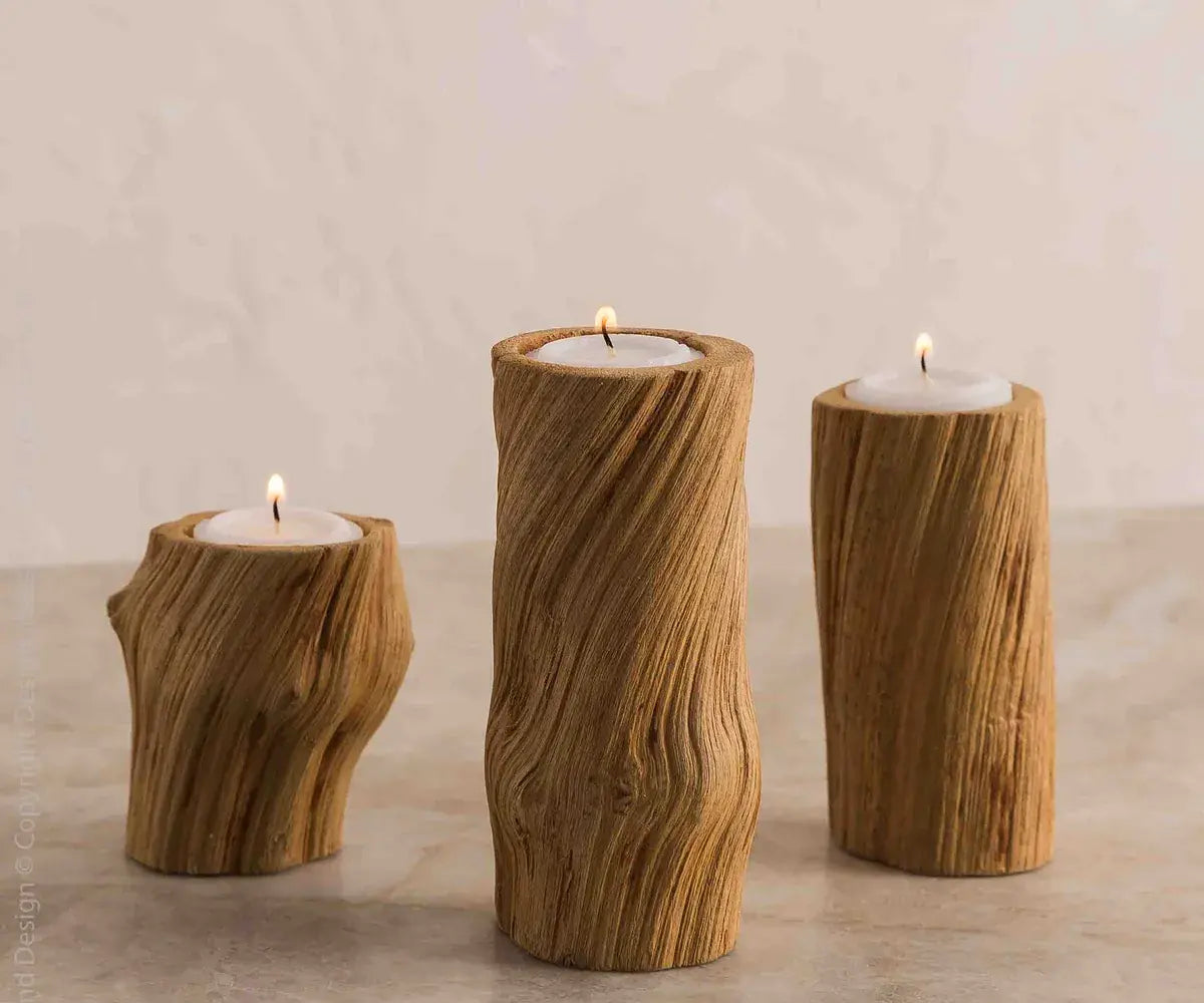 Three rustic wooden candle holders shaped like natural tree trunks, each holding a white tea light candle with a small flame, set against a neutral beige background.